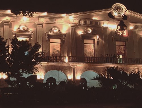 Mexiko Mérida - Palast Aufnahme am Abend. Der Palast ist schön beleuchtet. Hinter den Fenstern sieht man die ausgestellten Gemälde. Menschen stehen auf dem Balkon und schauen auf den Platz vor dem Palast. Vor dem Palast stehen Bäume, Sträucher und Palmen.