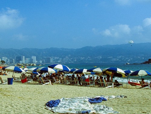 Mexiko Acapulco - Strand mit Liegen und Sonnenschirmen. Viele Menschen tummeln sich im Sand. Im Hintergrund Berge und Wolkenkratzer.