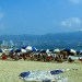 Mexiko Acapulco - Strand mit Liegen und Sonnenschirmen. Viele Menschen tummeln sich im Sand. Im Hintergrund Berge und Wolkenkratzer.
