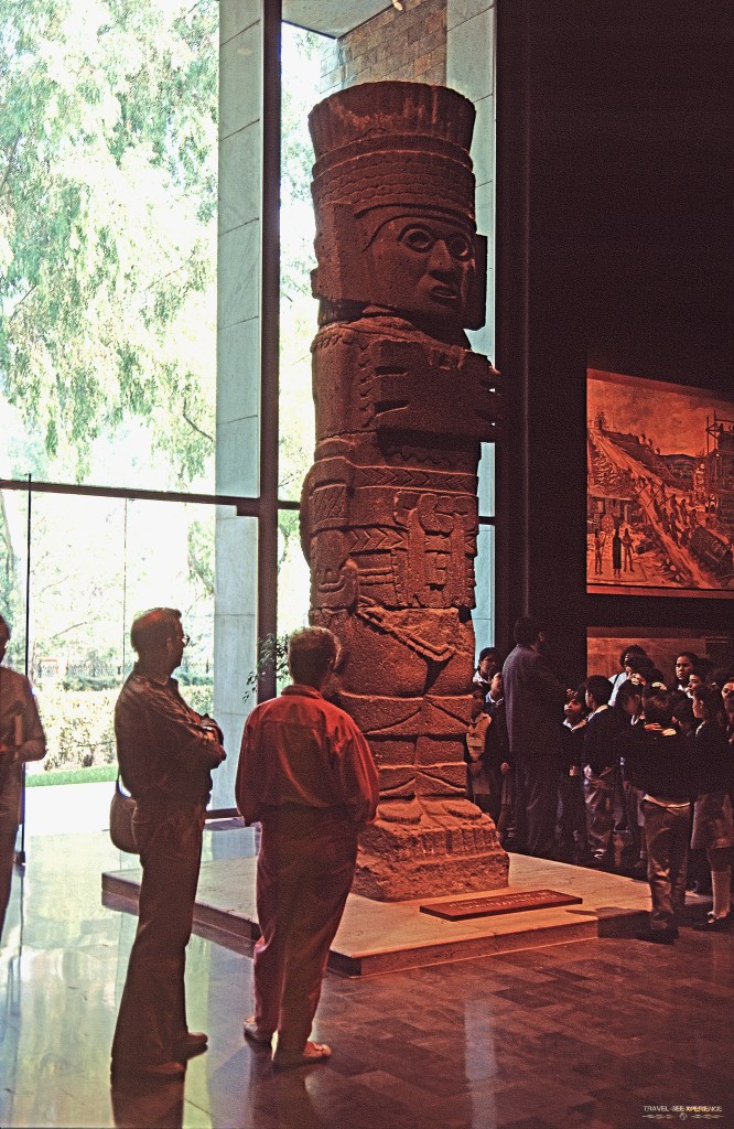Mexiko City - Eine riesige Säule mit einem filigran gestalteten Gesicht. Auch die Kleidung und der Kopfschmuck sind sehr gut ausgearbeitet. Die Stele ist aus Stein und stammt von den Tula Giants. Um sie herum stehen Menschen und betrachten sie. Die Säule steht vor einem großen Fenster.
