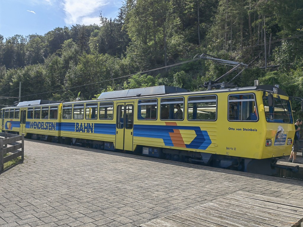 Wendelstein - Eine Wendelstein Zahnradbahn wartet an der Haltestelle, um Passagiere in die Berge zu bringen. Die gelb-blau gestrichene Bahn steht vor einer bewaldeten Kulisse.